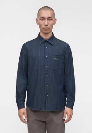 Homme portant une chemise bleu foncé à manches longues avec une poche poitrine et un pantalon marron clair, debout devant un fond blanc uni.