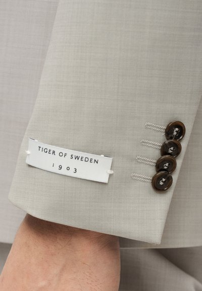 Μανίκι γκρι σακακιού με τέσσερα σκούρα κουμπιά, λευκή ετικέτα με το κείμενο «TIGER OF SWEDEN 1903» και διακριτή υφή.
