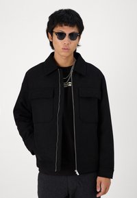 Veste noire à fermeture éclair avec un col pointu, deux poches avant et une texture douce. Portée par-dessus une chemise noire et complétée par des lunettes de soleil.