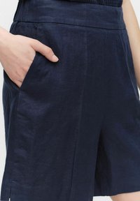 Tummansiniset pellavashortsit, joissa on sivutaskut, erottuvat sileästä tekstuurista ja puhtaista linjoista. Korostettu muotoilu on mukautettu vyötärölle ja yksinkertainen design.