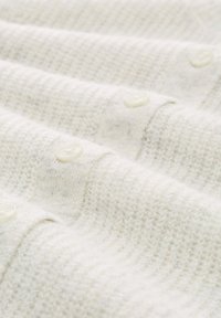 Tissu de couleur crème clair avec une texture en maille, présentant des boutons blancs le long du bord. Surface douce et légèrement duveteuse.