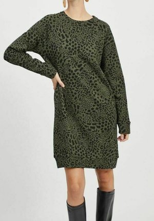 Vrouw draagt een olijfgroene knie-lange jurk met zwarte luipaardprint en lange mouwen, gecombineerd met zwarte kniehoge laarzen.