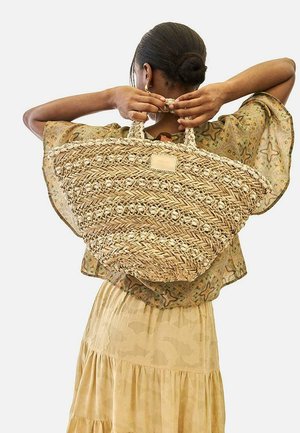 Mujer con moño trenzado sostiene un gran bolso de paja tejido detrás de la espalda, lleva una blusa transparente con estampado y una falda beige de varias capas.