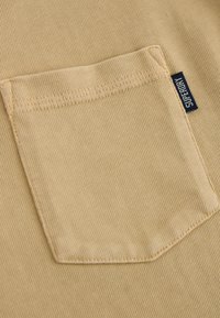 Tan tyg med en strukturerad yta har en patchficka. En liten, blå etikett med texten "SUPERDRY" är synlig nära fickan.