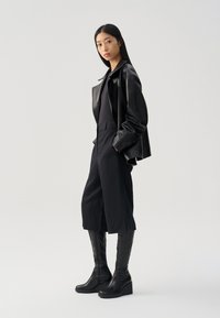 Chaqueta de cuero negra sobre una blusa negra ajustada, combinada con pantalones oscuros anchos y botas de cuña negras hasta la rodilla. Texturas suaves y diseño minimalista.