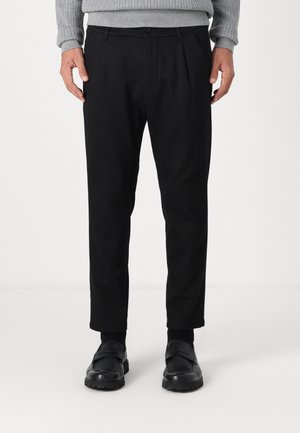 Uomo che indossa pantaloni neri su misura, mocassini neri, calze nere e un maglione grigio lavorato a maglia, in piedi davanti a uno sfondo semplice.