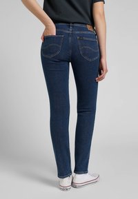 Skinny jeans in dark blue denim, featuring a mid-rise waist, back pockets with stitching detail, and a smooth texture. Worn with white sneakers.