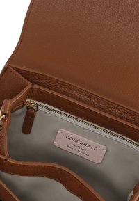 Interno di una borsa in pelle marrone con fodera grigio chiaro, dotata di tasca con zip e di un'etichetta che recita "Coccinelle Authentic Italian Leather."