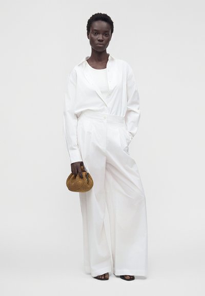 Femme portant un pantalon blanc à jambes larges et une chemise blanche boutonnée, tenant un petit sac à main tressé marron, debout devant un fond uni.