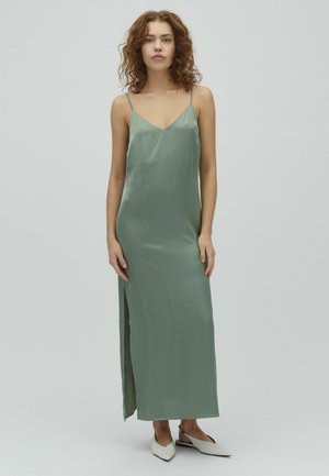 Kobieta o kręconych włosach, ubrana w bezrękawnikową, szałwiowozieloną suknię maxi z bocznym rozcięciem oraz białe baleriny typu slingback z ostrym noskiem na jednolitym tle.