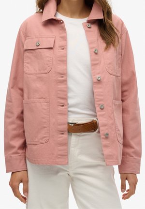 Persona che indossa una giacca rosa chiaro con bottoni e grandi tasche sopra una camicia bianca e pantaloni bianchi con cintura marrone.