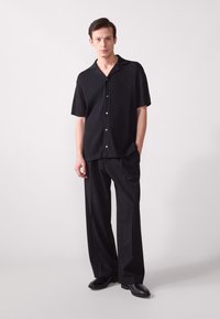 Chemise noire à manches courtes côtelée avec une fermeture à boutons, associée à un pantalon noir ample et des chaussures noires, sur un fond neutre.