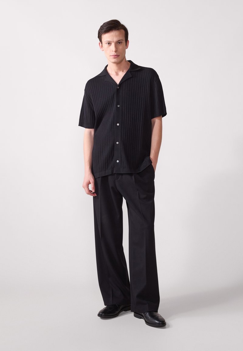 Chemise noire à manches courtes côtelée avec une fermeture à boutons, associée à un pantalon noir ample et des chaussures noires, sur un fond neutre.