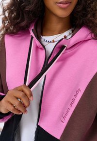 Bluza z kapturem w kolorze różowym, brązowym i czarnym, z zamkiem z przodu. Posiada mały haftowany napis "Wieczność zaczyna się dzisiaj" na różowej części.