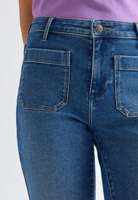 Gros plan de la section médiane portant un jean en denim bleu taille haute avec des poches plaquées à l’avant et un haut violet rentré.