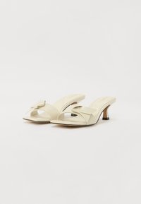 Beige high-heeled sandals with a wide strap, an oversized bow detail, open toe, and a short, black-accented heel. Smooth texture.