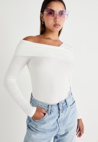 Witte off-shoulder top met lange mouwen, gecombineerd met lichtblauwe high-waisted jeans. Het model draagt overgrote roze zonnebrillen. Gladde textuur.