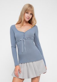Top albastru cu mâneci lungi, decolteu în formă de rotund, butoane decorative, dantelă pe margini și detaliu cu șnur la talie, asortat cu o fustă gri plisată.