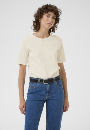 Femme aux cheveux bouclés portant une chemise crème à manches courtes, un jean bleu et une ceinture noire, debout devant un fond clair uni.