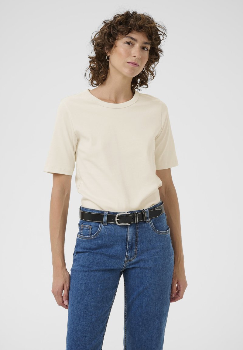 Femme aux cheveux bouclés portant une chemise crème à manches courtes, un jean bleu et une ceinture noire, debout devant un fond clair uni.