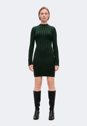 Un mannequin se tient face à l’objectif, vêtu d’une robe côtelée vert foncé à manches longues et de bottes noires montant jusqu’aux genoux, sur un fond clair uni.