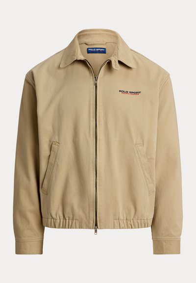 Μπεζ σακάκι με γιακά, εμπρόσθιο φερμουάρ και ελαστικά μανίκια. Διαθέτει πλαϊνές τσέπες και λογότυπο "Polo Sport" σε κόκκινο και μπλε.
