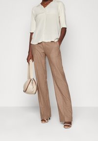 Blouse en crème clair avec un col en V et des manches trois-quarts, assortie à un pantalon large à carreaux marron ; porte un sac beige à finition texturée.