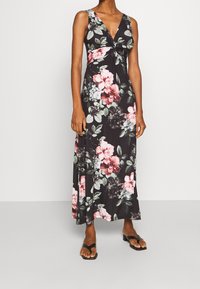 Robe longue fleurie en tissu noir, dotée d'un décolleté en V profond, d'une taille nouée et de grands motifs floraux roses et blancs. Sandales plates noires.