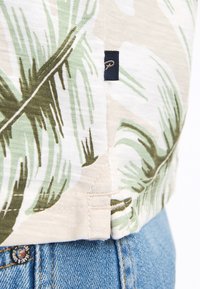 Camicetta a maniche corte con una base beige e stampa di foglie di palma verdi. Cucitura laterale visibile con un piccolo etichetta blu navy. Realizzata in tessuto leggero.