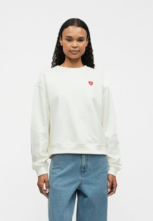 Ljus krämfärgad sweatshirt med ribbade mudd och fåll, prydd med en liten röd hjärt-logotyp på vänster bröst. Bärs tillsammans med blå denimjeans.
