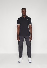 Čierna polo tričko s bielymi a zlatými akcentmi na golieri, skombinované s tmavými, ľahkými nohavicami. Model má obuté biele športové topánky.