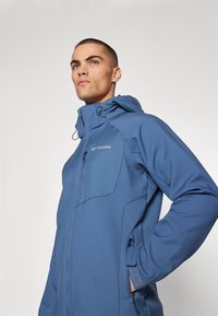 Niebieska kurtka typu softshell z zamkiem z przodu, regulowanym kapturem i bocznymi kieszeniami. Posiada gładką teksturę oraz kontrastowe szwy.
