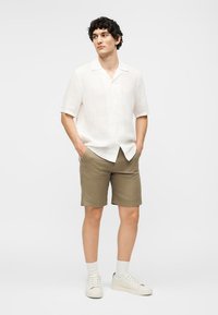 Άνδρας που στέκεται με τα χέρια στις τσέπες, φοράει λευκό πουκάμισο με κοντά μανίκια και κουμπιά, ελιά σορτς, λευκές κάλτσες και λευκά αθλητικά παπούτσια σε λευκό φόντο.