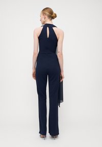 Macacão azul-marinho com decote halter e costas em forma de chave. Apresenta um corte ajustado e pernas largas, complementado por um detalhe de tecido transparente nas costas.