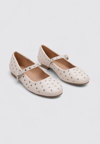 Zapatos planos beige con puntera redonda, con una correa en el empeine y tachuelas metálicas en la parte superior. Textura de cuero suave, tacón bajo.