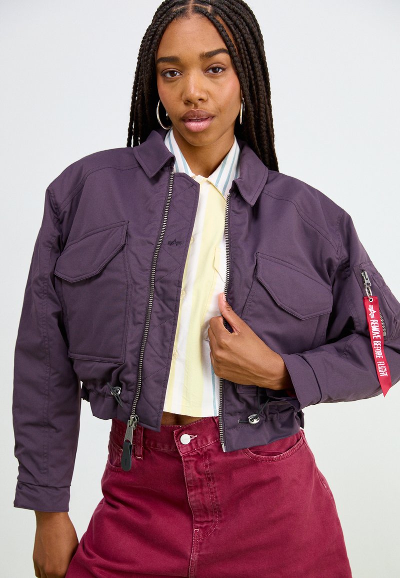 Mujer con trenzas largas que lleva una chaqueta bomber púrpura corta sobre una camisa a rayas amarillas y pantalones rojos, sujetando la cremallera de la chaqueta con una mano.