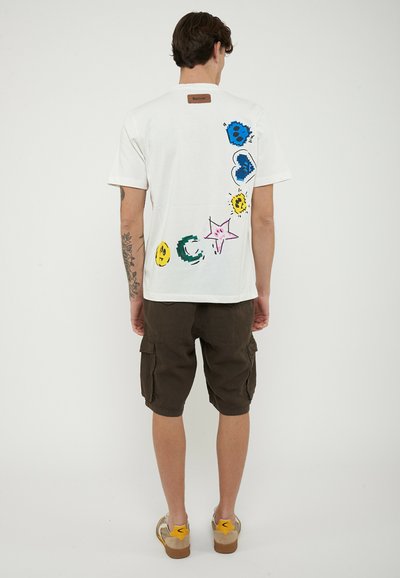 Uomo che indossa una t-shirt bianca con forme astratte colorate sulla schiena, pantaloncini cargo scuri e sneakers beige con dettagli gialli, in piedi di spalle.