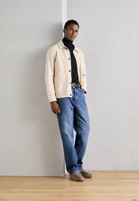 Veste en coton beige avec poches avant, col roulé noir, jeans en denim bleu et chaussures grises avec semelles en caoutchouc, se tenant contre un mur.