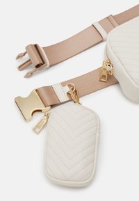 Křížová taška z umělé kůže v bílé barvě s chevronovým vzorem, zlatými kovovými doplňky a odnímatelným béžovým páskem, vybavená malou kapsičkou na zip.