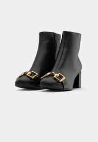Bottines noires en cuir avec bouts ouverts, fermeture éclair sur le côté et accents en boucle dorée, dotées d'un talon épais et d'une texture lisse.