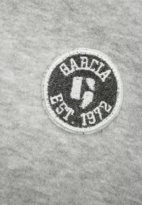 Šedá tkanina s výšivkou černobílého loga v kruhovém tvaru, které obsahuje text "GARCÍA EST. 1972". Logo je vyšíváno s texturovanými detaily.