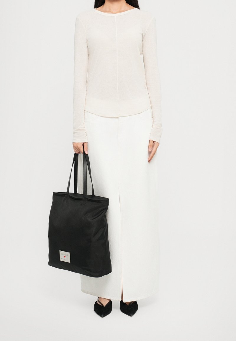 Femme portant une longue jupe blanche et un haut à manches longues, tenant un grand sac cabas noir avec des bretelles d'épaule sur fond blanc.