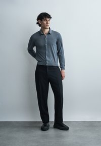 Homme aux cheveux bouclés portant un cardigan gris boutonné, un pantalon noir et des chaussures noires, debout contre un mur gris clair uni.