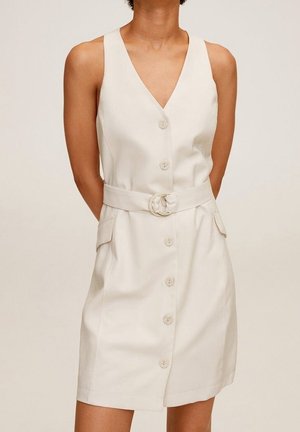 Biała, bezrękawnikowa sukienka z przodu na guziki, dekoltem w serek, paskiem w talii i kieszeniami z klapkami, noszona przez osobę stojącą z rękami za plecami.