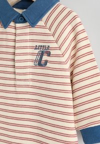 Polo w paski w kolorze kremowym z czerwonymi paskami i niebieskim kołnierzykiem. Zawiera haftowane logo "Little C". Miękka tekstura z rękawami raglanowymi.