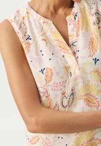 Blouse sans manches en coton doux avec un fond crème, présentant des motifs floraux colorés en rose, jaune et bleu, et une patte de boutonnage partielle.