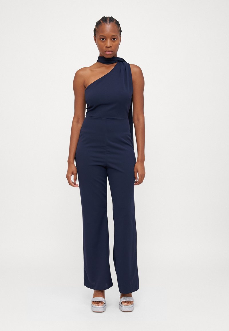 Macacão azul-marinho de um ombro só, com corpete ajustado e pernas largas, apresentando um detalhe drapeado no ombro e uma textura de tecido suave.