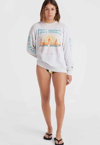 Felpa grigia con grafiche stampate di sole e figure, maniche lunghe, polsini e orlo a costine, indossata con un bikini giallo e nero.