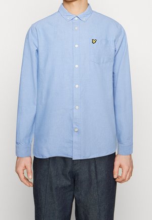 Mann trägt hellblaues Hemd mit Knöpfen und Kragen sowie gelbem Bienen-Logo auf der linken Brusttasche, kombiniert mit dunkler Hose.