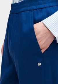 Άτομο που φοράει μπλε παντελόνι με ελαστική μέση και έχει το χέρι στην τσέπη, συνδυασμένο με μπλε και λευκό ριγέ πουκάμισο με μανίκι που έχει γυρισμένο μανσέτο.